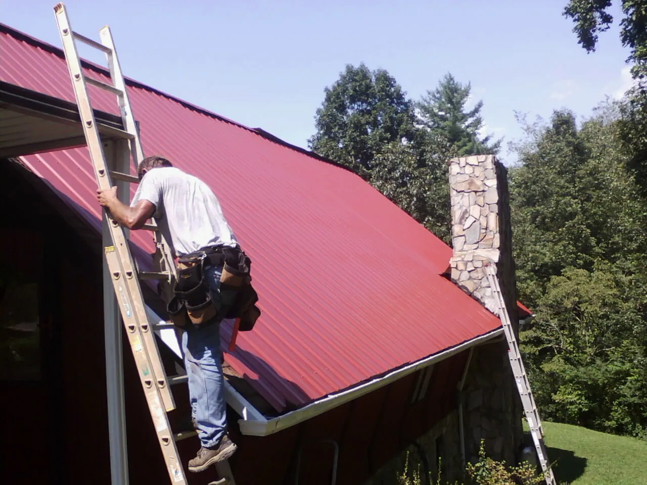 Roofer on a ladder beside a red metal roof and stone chimney — Soffit & Fascia Repair by Roofing Brothers in Florahome